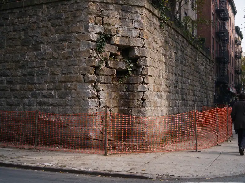 Typical condition: a tall mortared stone retaining wall bulging with missing stones, fenced off from the sidewalk in the Bronx