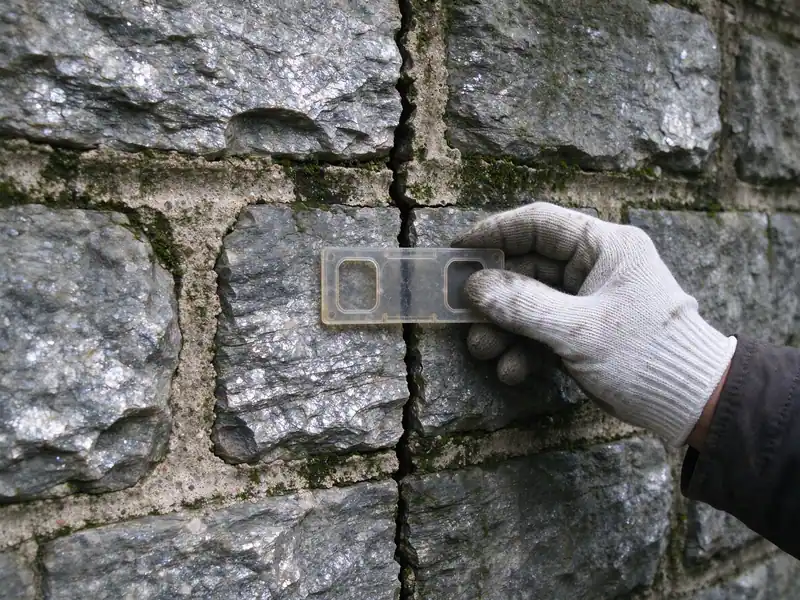 Gloved hand holding a crack-width gauge across a mortar crack in a Manhattan schist retaining wall in Inwood